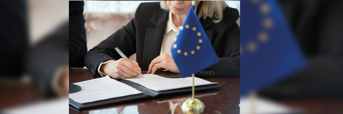 Mano firma documento oficial en mesa con banderín de la UE en primer plano y carpeta debajo.