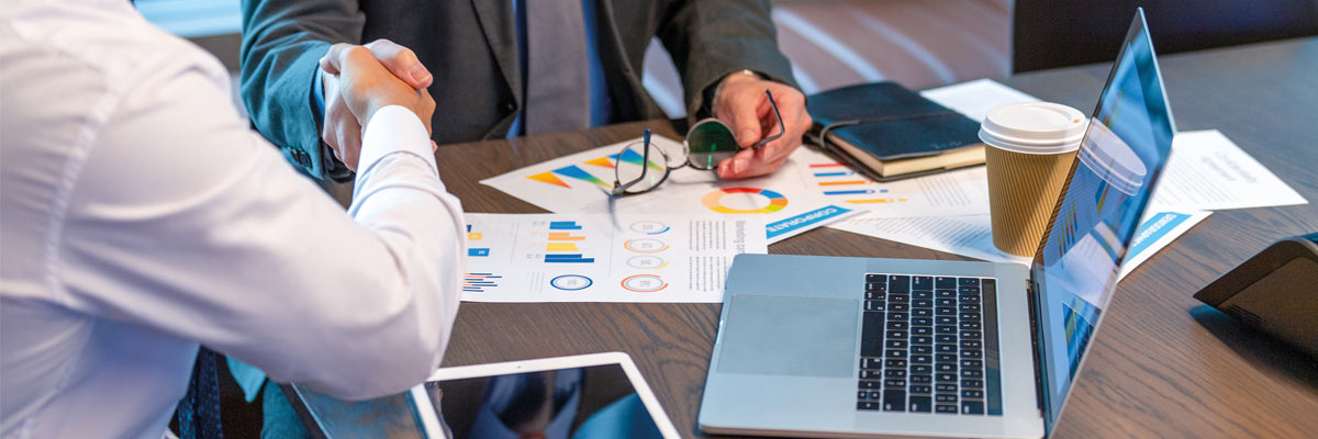 Dos personas en una mesa con documentos, gráficos, una laptop, tabletas digitales y una taza de café desechable, representando un entorno de reunión corporativa.