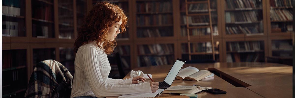 Estudiante universitaria estudia en biblioteca con portátil y apuntes, concentración y estudio académico.