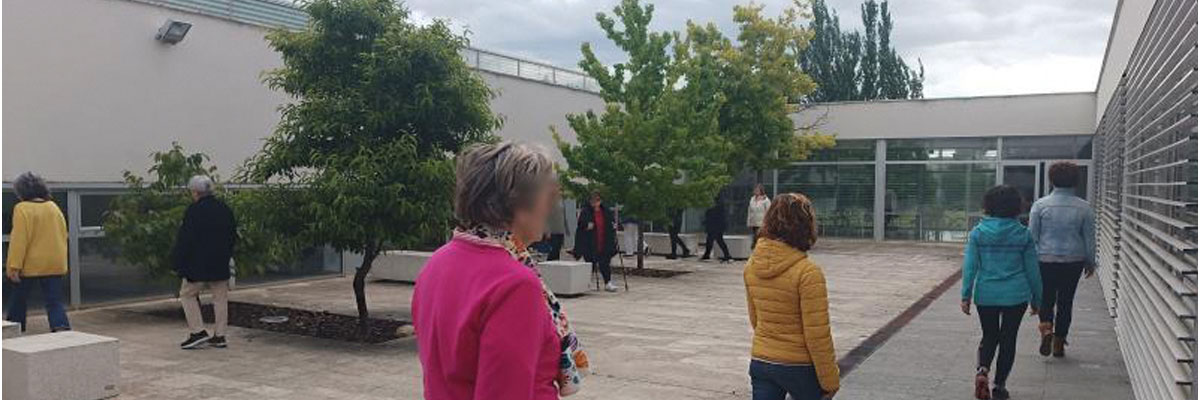 grupo de personas caminando en un entorno exterior, que parece ser una terraza o patio. Hay árboles y bancos, y el cielo está nublado. 