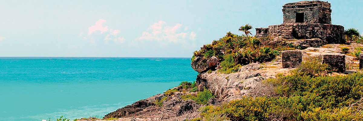 La imagen muestra un paisaje costero con cielos despejados y el mar. A la derecha, hay una estructura de piedra de la antigua Maya