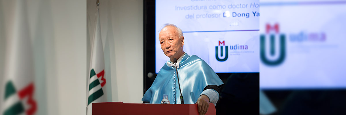 Dong Yansheng en una ceremonia de investidura, vistiendo una toga azul y de pie detrás de un podio, con banderas y un proyector mostrando texto en el fondo, capturando un logro académico significativo