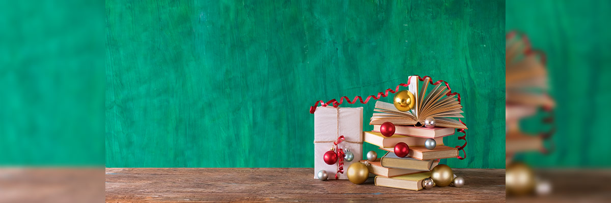 Una pila de libros adornada con esferas navideñas y una cinta roja en una mesa de madera, con un fondo verde