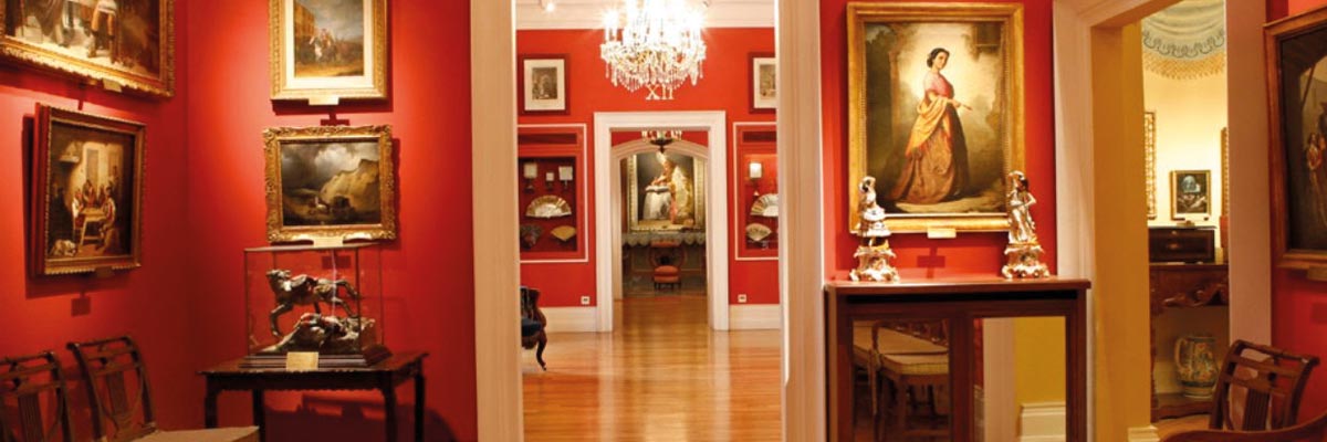 Sala de museo con paredes rojas, cuadros dorados y esculturas en vitrinas, suelo de madera y lámpara de araña colgante