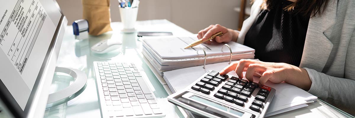 Mano de persona calculando con calculadora sobre mesa de cristal, junto a carpetas, bolígrafo y monitor con datos financieros