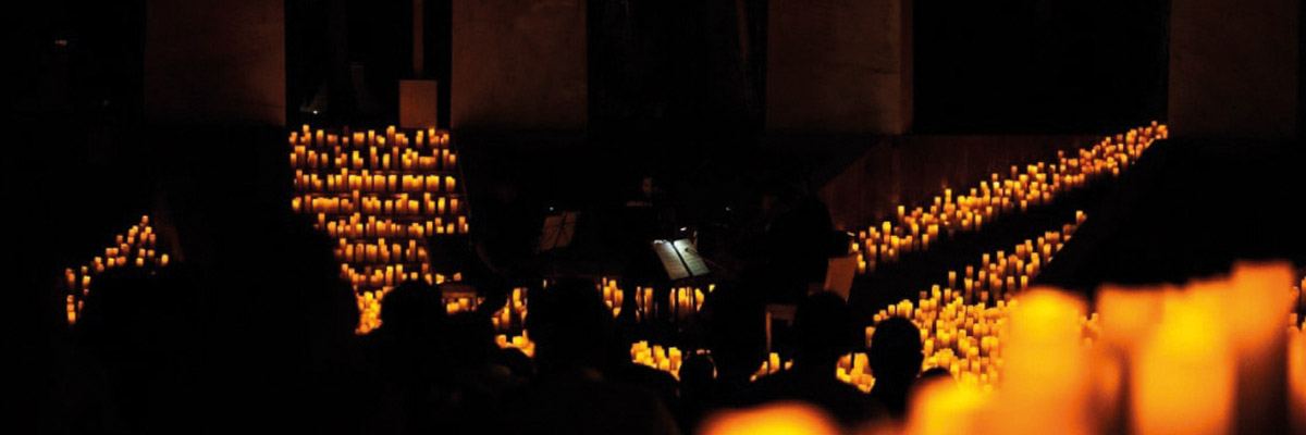 Sala oscura iluminada por cientos de velas dispuestas en escaleras, músicos tocando en penumbra