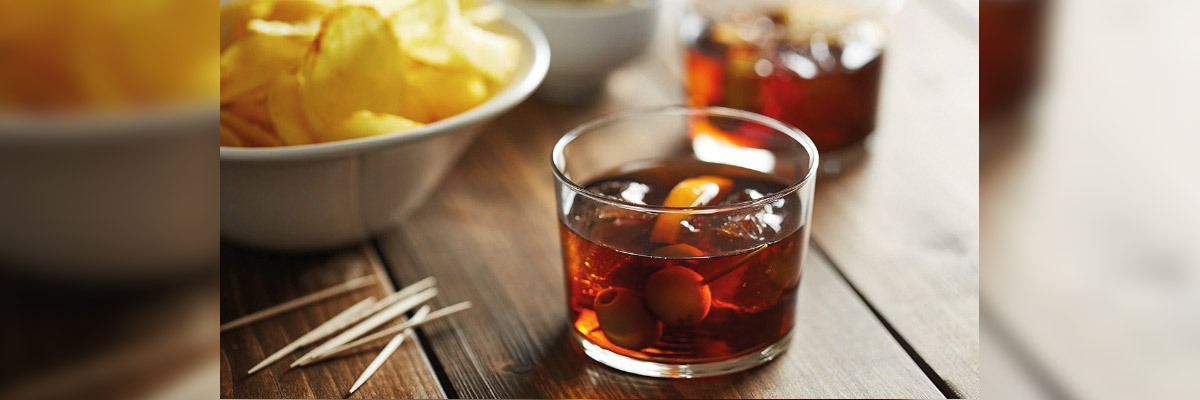 Vaso de vermut con aceitunas en primer plano, bol de patatas fritas y palillos sobre mesa de madera.