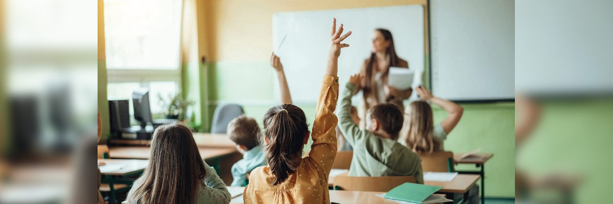 Aula con varios niños levantando la mano mientras una maestra imparte la clase con pizarra y pupitres al fondo