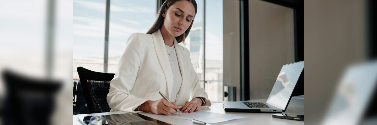 Mujer de negocios sentada en un escritorio moderno, escribiendo en un documento con un portátil abierto a su lado