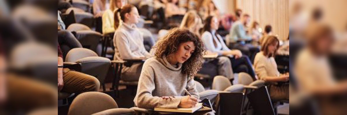 Estudiantes en un aula tipo anfiteatro, varios tomando apuntes, una joven al frente concentrada escribiendo en su libreta.