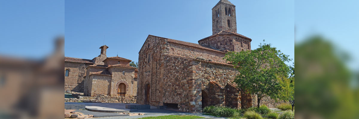 Edificio románico con torre de campanario y muros de piedra, rodeado de césped y cielo azul