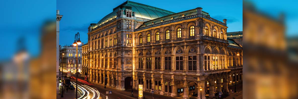 Fachada iluminada de la Ópera Estatal de Viena al anochecer, con tráfico y luces de ciudad alrededor