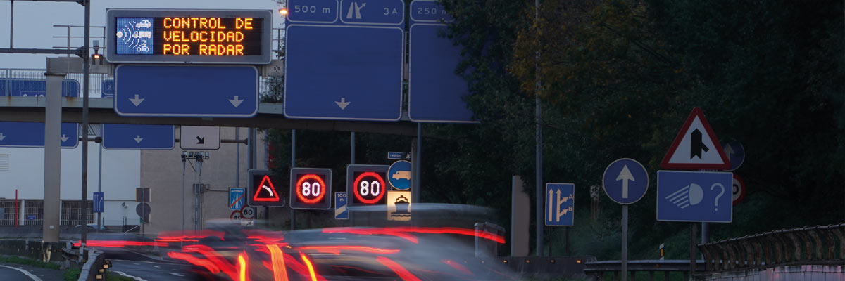 Señales de tráfico que indican control de velocidad por radar y límite de 80 km/h, con luces traseras de vehículos en movimiento al anochecer.