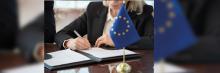 Mano firma documento oficial en mesa con banderín de la UE en primer plano y carpeta debajo.