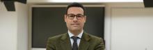 Retrato de Miguel Ortego, con gafas y chaqueta, de pie frente a una pizarra y pantalla en un entorno de aula