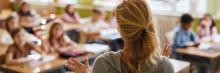 Profesora frente a alumnos en aula; estudiantes levantan la mano participando activamente en la clase