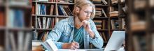 Joven rubio con gafas estudiando en biblioteca, escribe en un cuaderno frente a un portátil