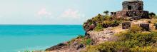 La imagen muestra un paisaje costero con cielos despejados y el mar. A la derecha, hay una estructura de piedra de la antigua Maya