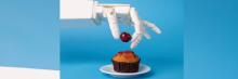 Mano robótica sujetando una cereza sobre un cupcake con glaseado, fondo azul liso.