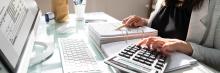 Mano de persona calculando con calculadora sobre mesa de cristal, junto a carpetas, bolígrafo y monitor con datos financieros