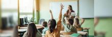 Aula con varios niños levantando la mano mientras una maestra imparte la clase con pizarra y pupitres al fondo