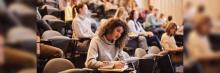 Estudiantes en un aula tipo anfiteatro, varios tomando apuntes, una joven al frente concentrada escribiendo en su libreta.